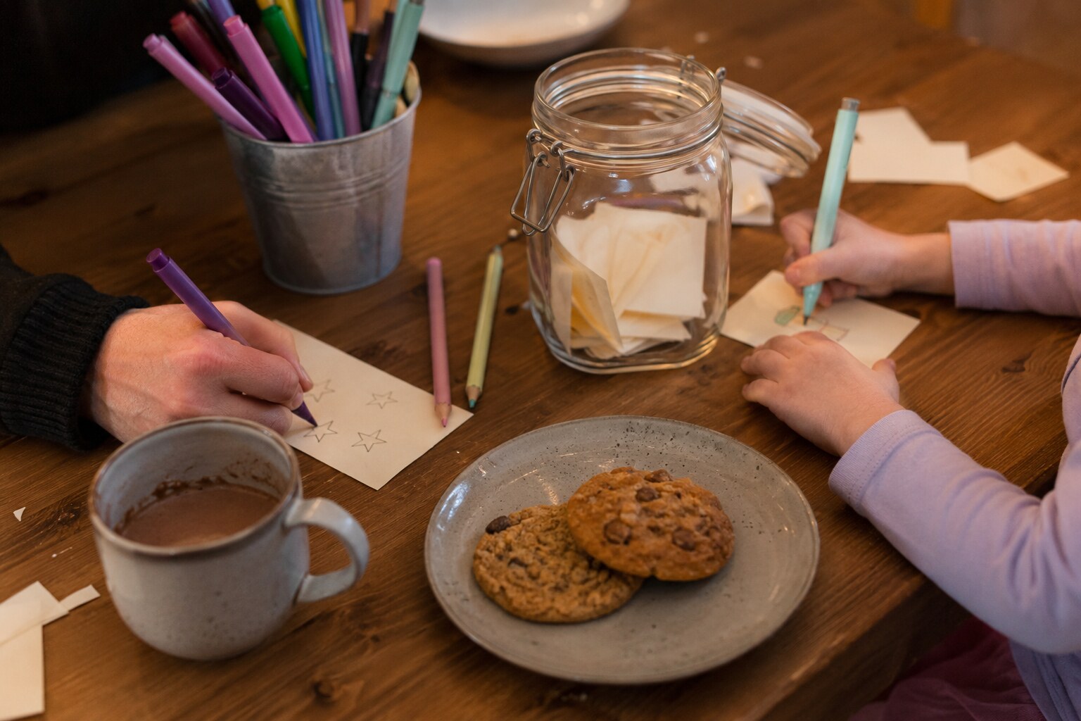 A parent and child making simple activity slips for a family tradition jar with markers, cocoa and cookies