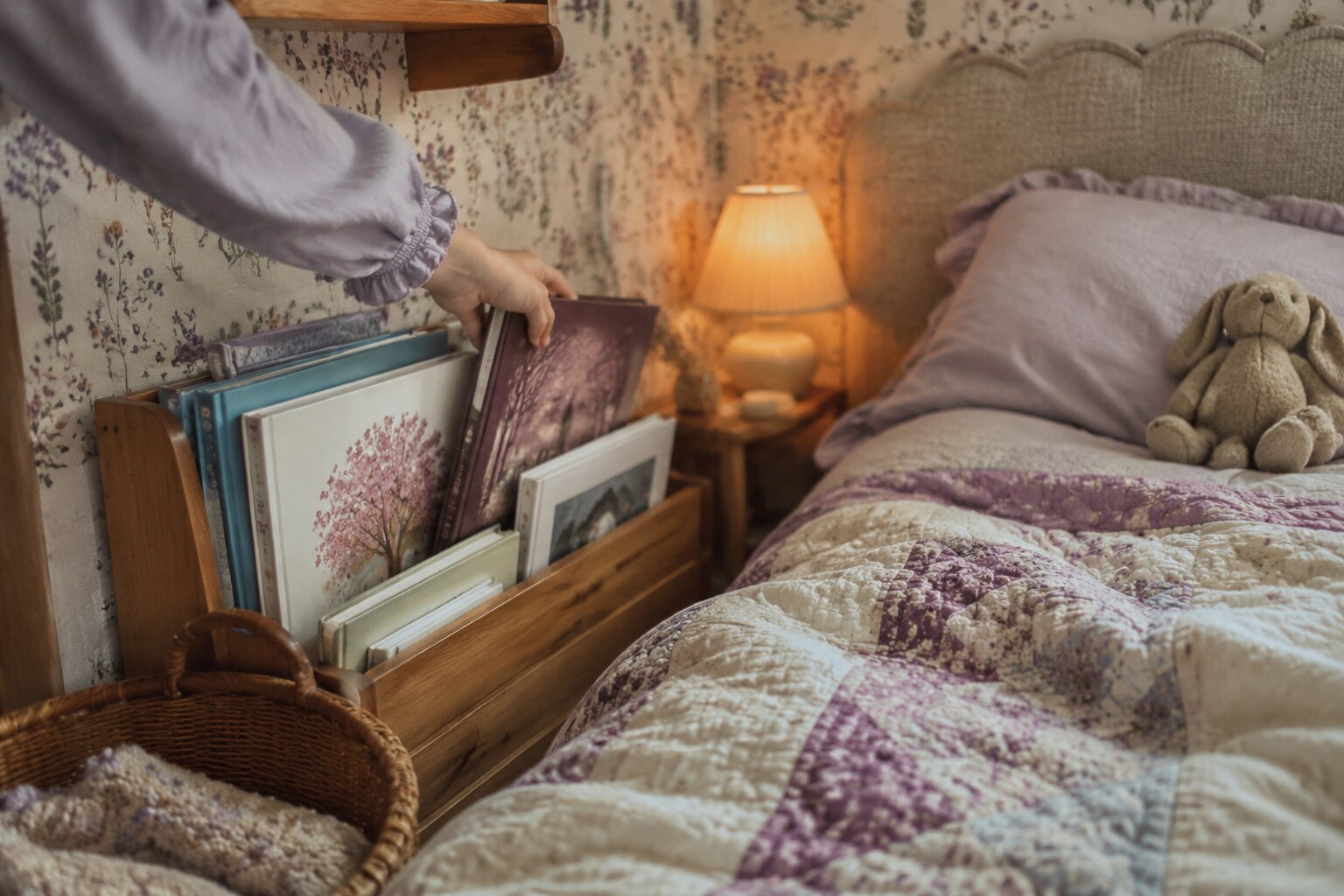 A child choosing a picture book from a low wooden shelf beside a floral quilt and warm reading lamp